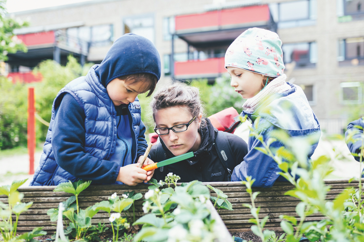 Ein Kind steht neben einem Hochbeet und untersucht mit einer Lupe Pflanzen. Ein anderes Kind und eine erwachsene Person schauen dabei zu