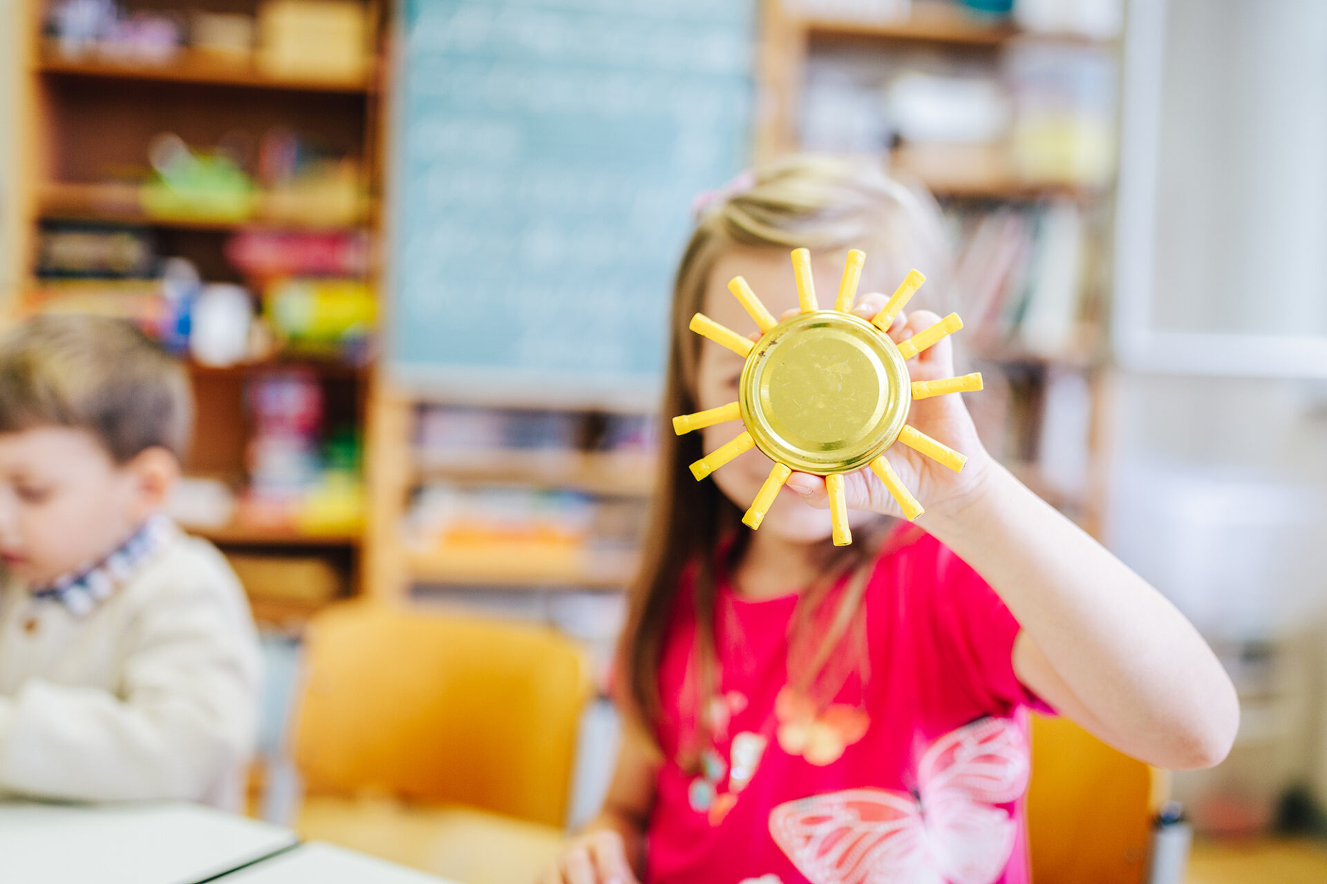 Ein Kind hält eine gebastelte Sonne aus einem Schraubdeckel und gelben Plastikteilen als Strahlen hoch